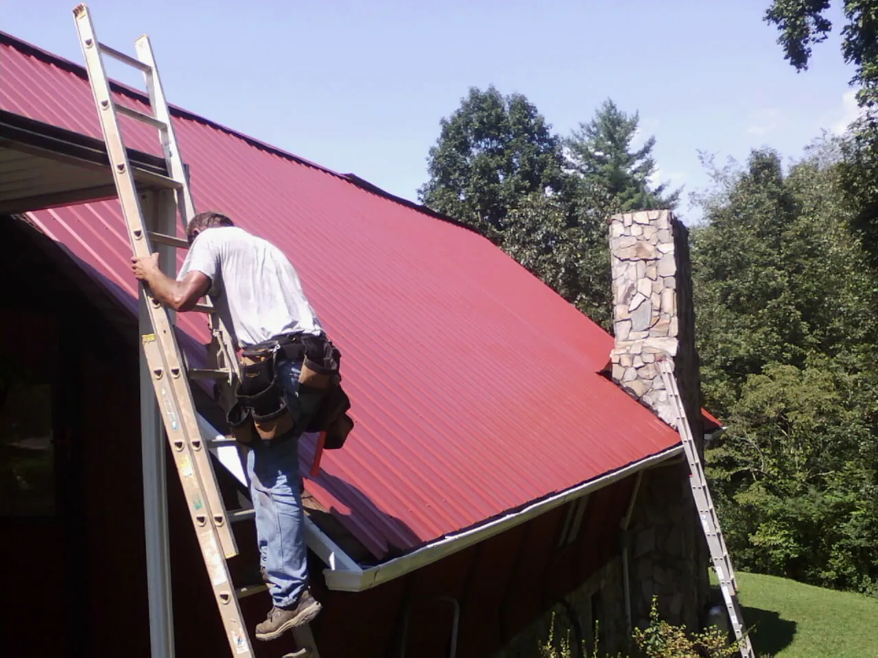 Roofer on a ladder beside a red metal roof and stone chimney — Soffit & Fascia Repair by Roofing Brothers in Chesterfield